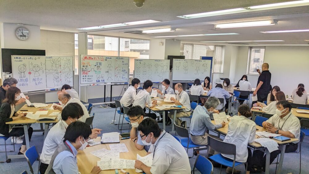 企業研修で、参加者がグループに分かれて、ホワイトボードの棒人間を見ながらイラストを描いている様子