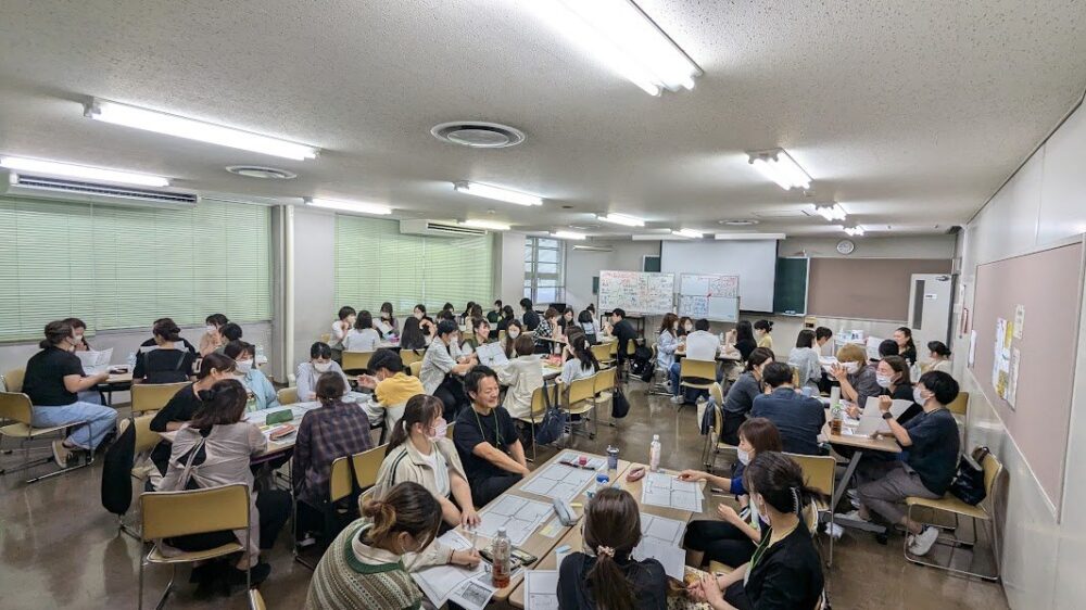 広い部屋で、多くの参加者がグループに分かれて、紙に描きながら話し合っている様子