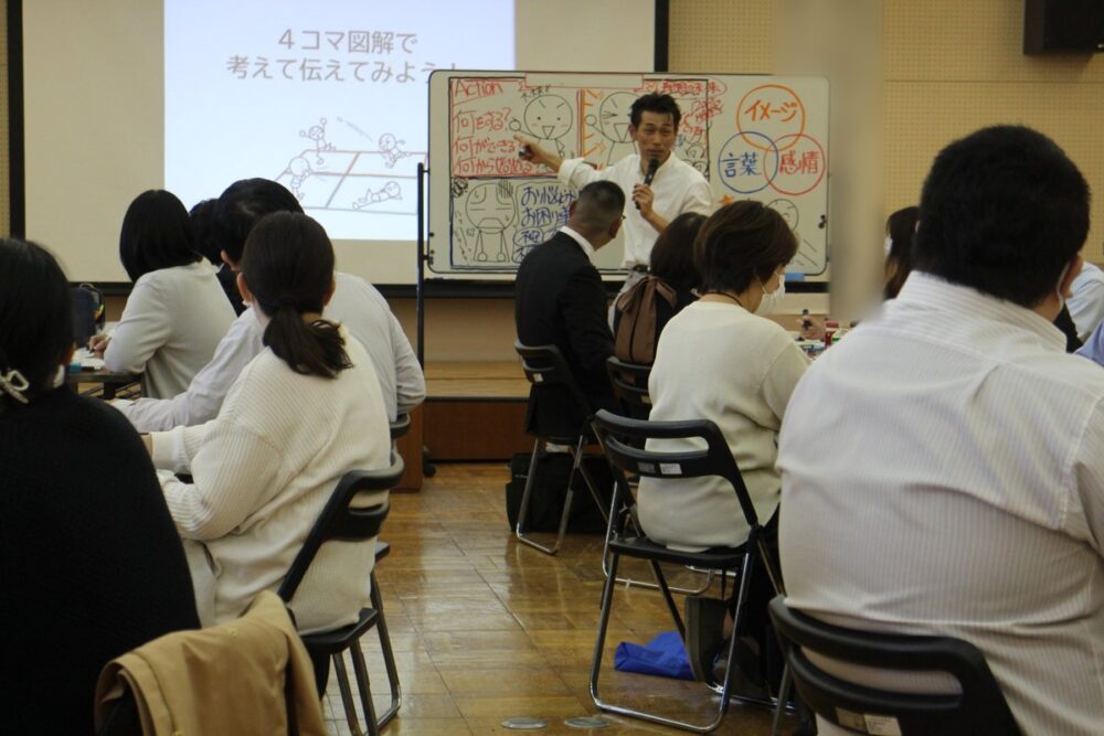 講師がマイクを持って、ホワイトボードの棒人間と図を指しながら、参加者に説明している様子