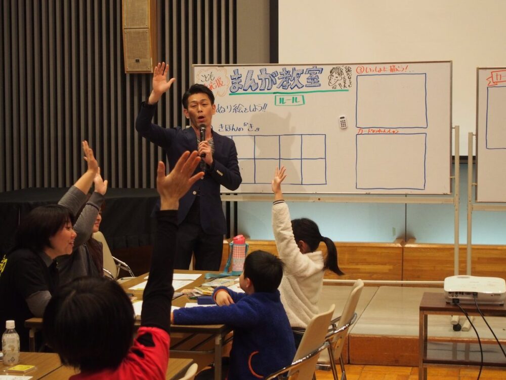 子ども向けのまんが教室で、講師の問いかけに、子どもと大人が元気に手を挙げている様子